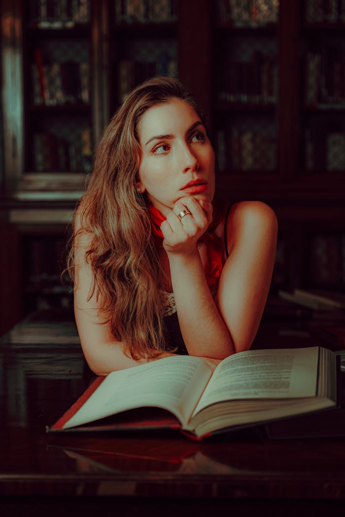 A contemplative woman sits in a library with an open book, deep in thought.